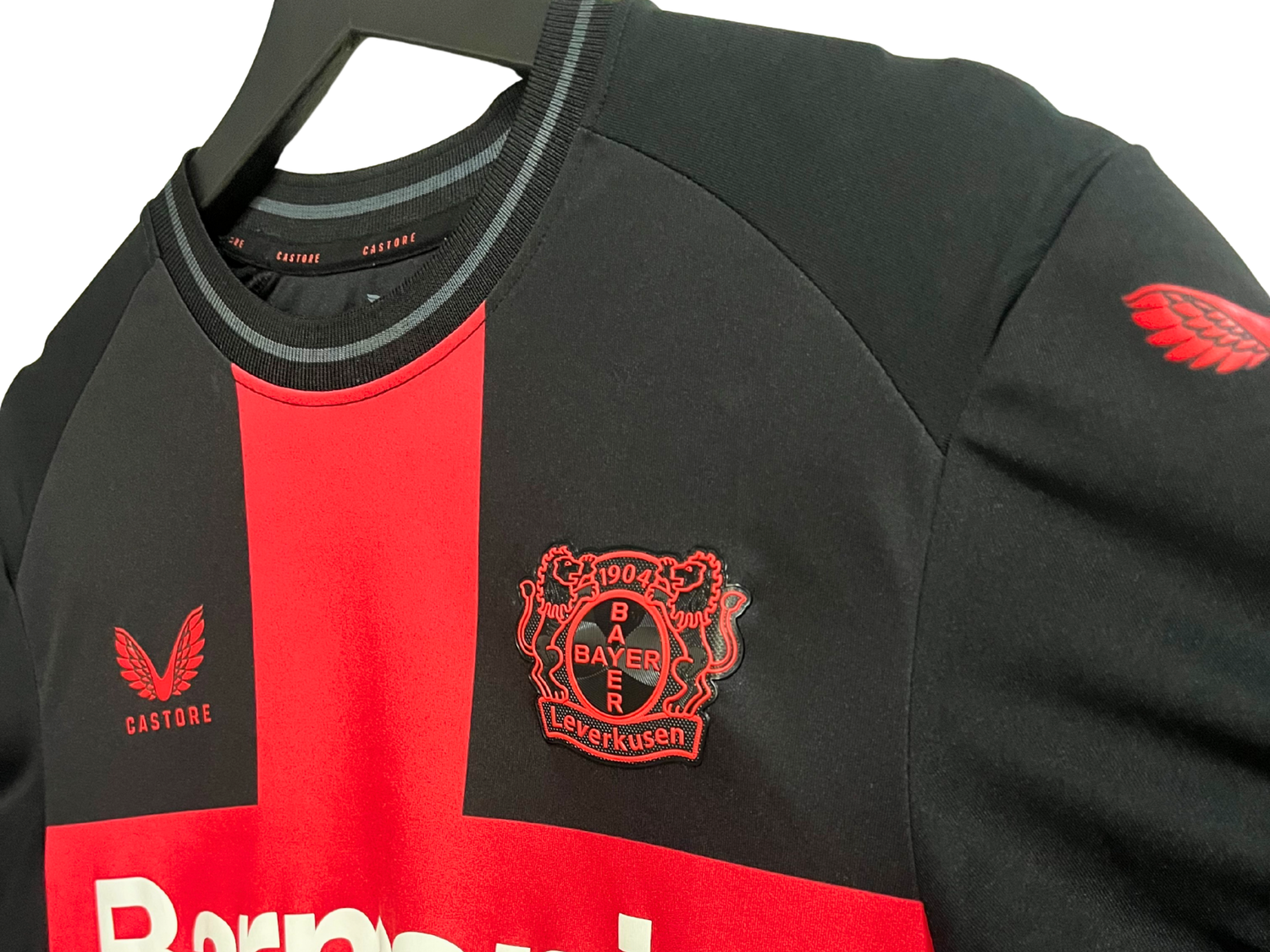 Détail du maillot Bayer Leverkusen VAPOR domicile 2023/2024 : gros plan sur la bande rouge avec logo du club et logo Castore.